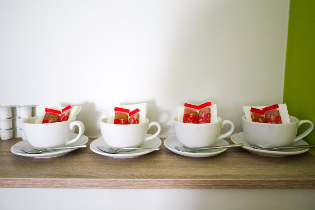 Four white teacups with saucers and spoons are arranged in a row on a wooden shelf, each containing sugar packets and napkins.