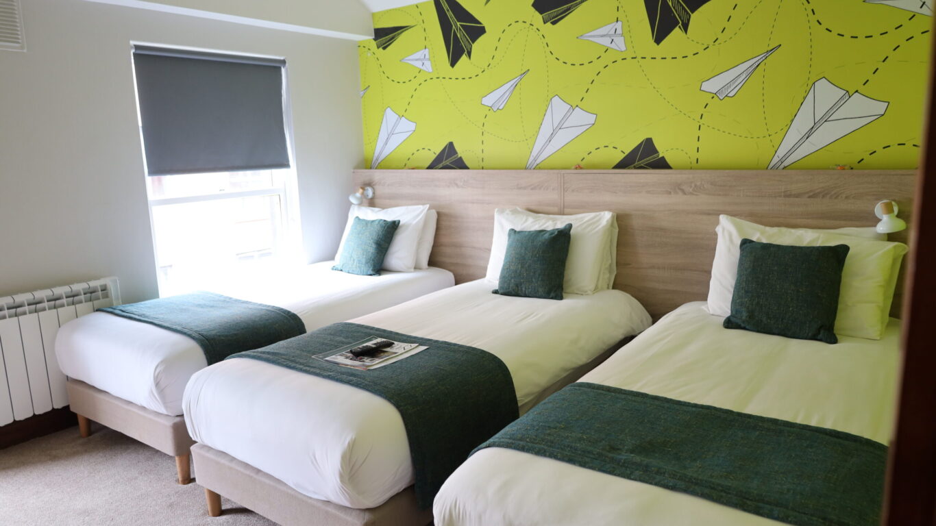 Three single beds with white linens and green throws are arranged in a row in a room with a window and a yellow wall featuring a paper airplane pattern.