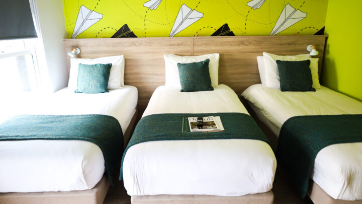 Three single beds with teal throws and pillows are arranged side by side in a brightly lit room with green, paper airplane patterned wallpaper. A magazine is on the middle bed.
