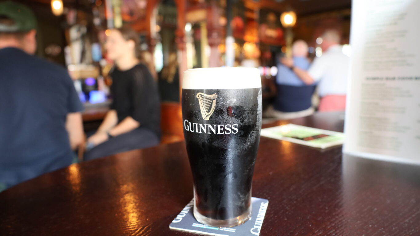 A pint of Guinness stout on a coaster sits on a wooden table in a pub, with people and bar decor visible in the background.