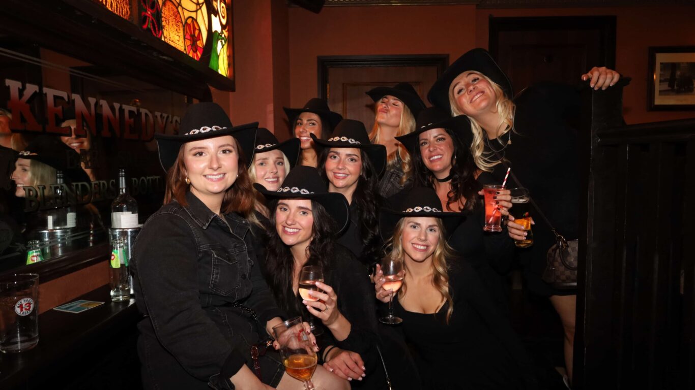 A group of women in black outfits and cowboy hats pose together indoors, some holding drinks and smiling at the camera.