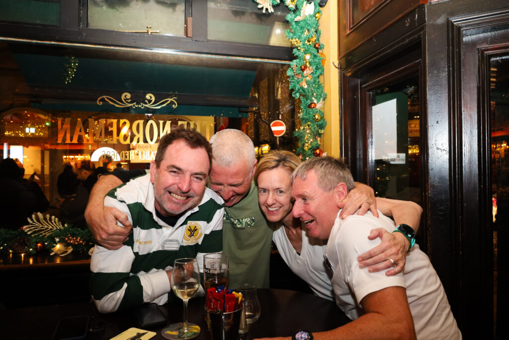 Four adults sit closely together at a bar table, smiling and embracing, with drinks and festive holiday decorations in the background.