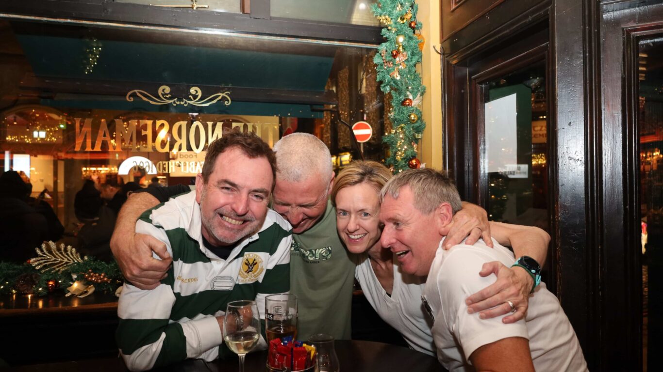 Four adults sit closely together at a pub table, smiling and hugging, with drinks and festive decorations visible in the background.
