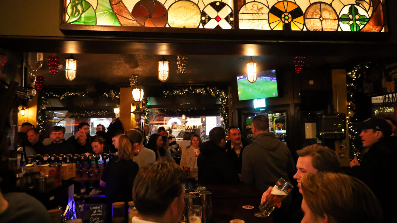 Crowded pub interior with people socializing at the bar, drinks in hand, and a sports game playing on a mounted TV. Stained glass decor and warm lighting set the atmosphere.