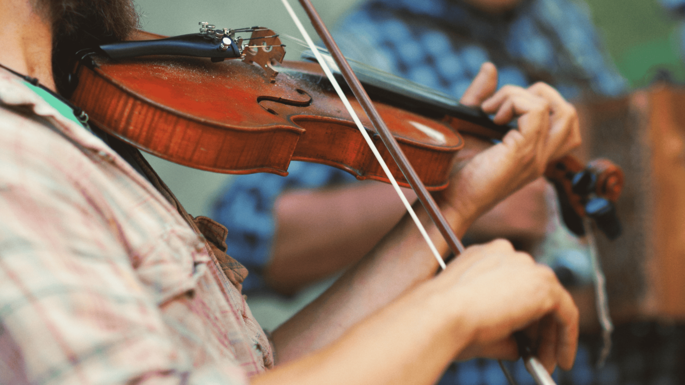 Close-up of a person playing a violin with a bow, wearing a plaid shirt. Another person in a blue plaid shirt is blurred in the background.