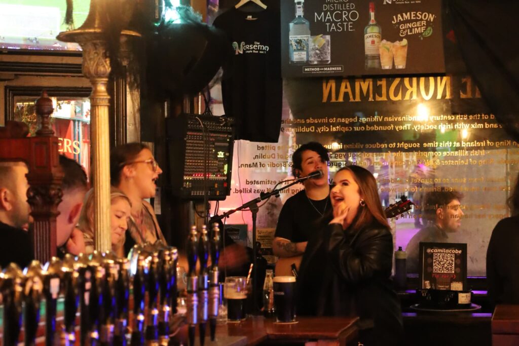 People are socializing and singing in a lively bar, with a musician performing in the background and beer taps visible in the foreground.