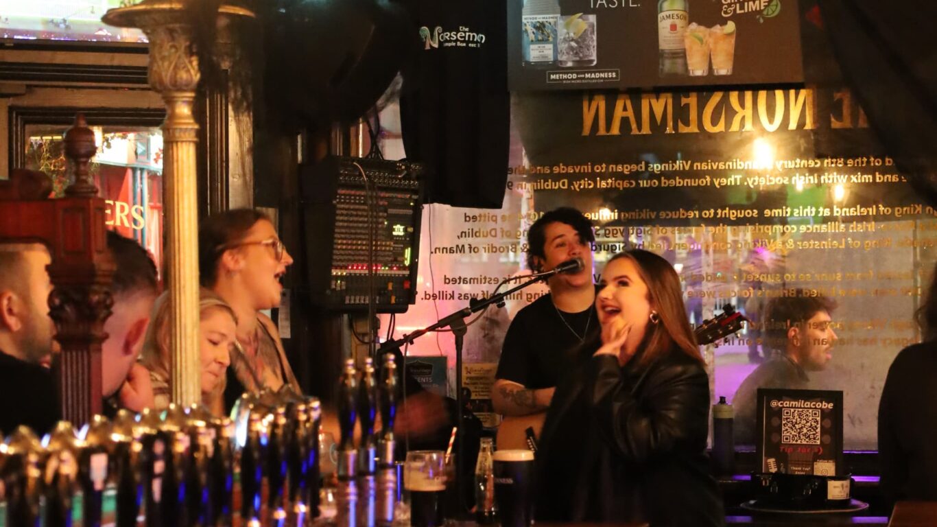 People are socializing and singing in a lively bar, with a musician performing in the background and beer taps visible in the foreground.