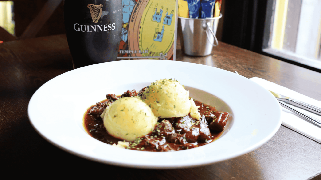 A plate of beef stew with two scoops of mashed potatoes, a pint of Guinness, and utensils on a wooden table near a window.