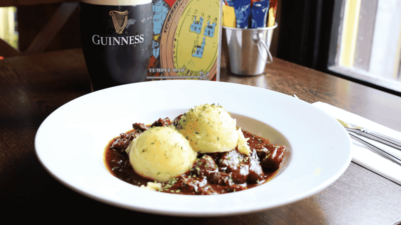 A plate of beef stew with two scoops of mashed potatoes, a pint of Guinness, and utensils on a wooden table near a window.