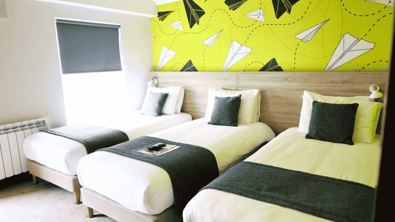 A hotel room with three single beds, each with gray accents, against a wall decorated with paper airplane illustrations on a yellow background.