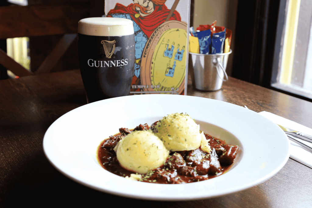 Untitled design - 2026-01-28T155903.926 A plate of beef stew with two scoops of mashed potatoes sits on a wooden table beside a pint of Guinness and a metal cup holding condiment packets.