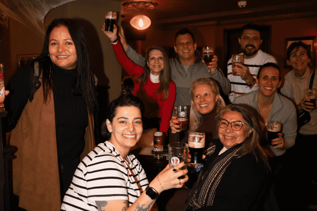 A group of people smile and raise glasses of beer while gathered around a table in a dimly lit bar or pub.