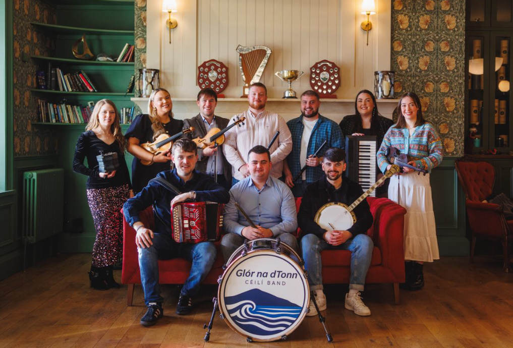 A group of ten musicians pose indoors with their instruments, including an accordion, banjo, keyboard, flute, and drum marked 
