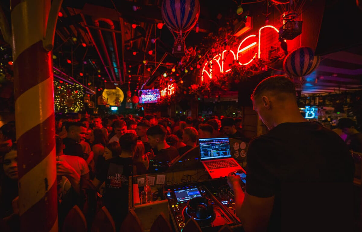 Vibrant hotel nightclub with lively crowd, DJ playing upbeat music, illuminated neon signs create an energetic atmosphere.